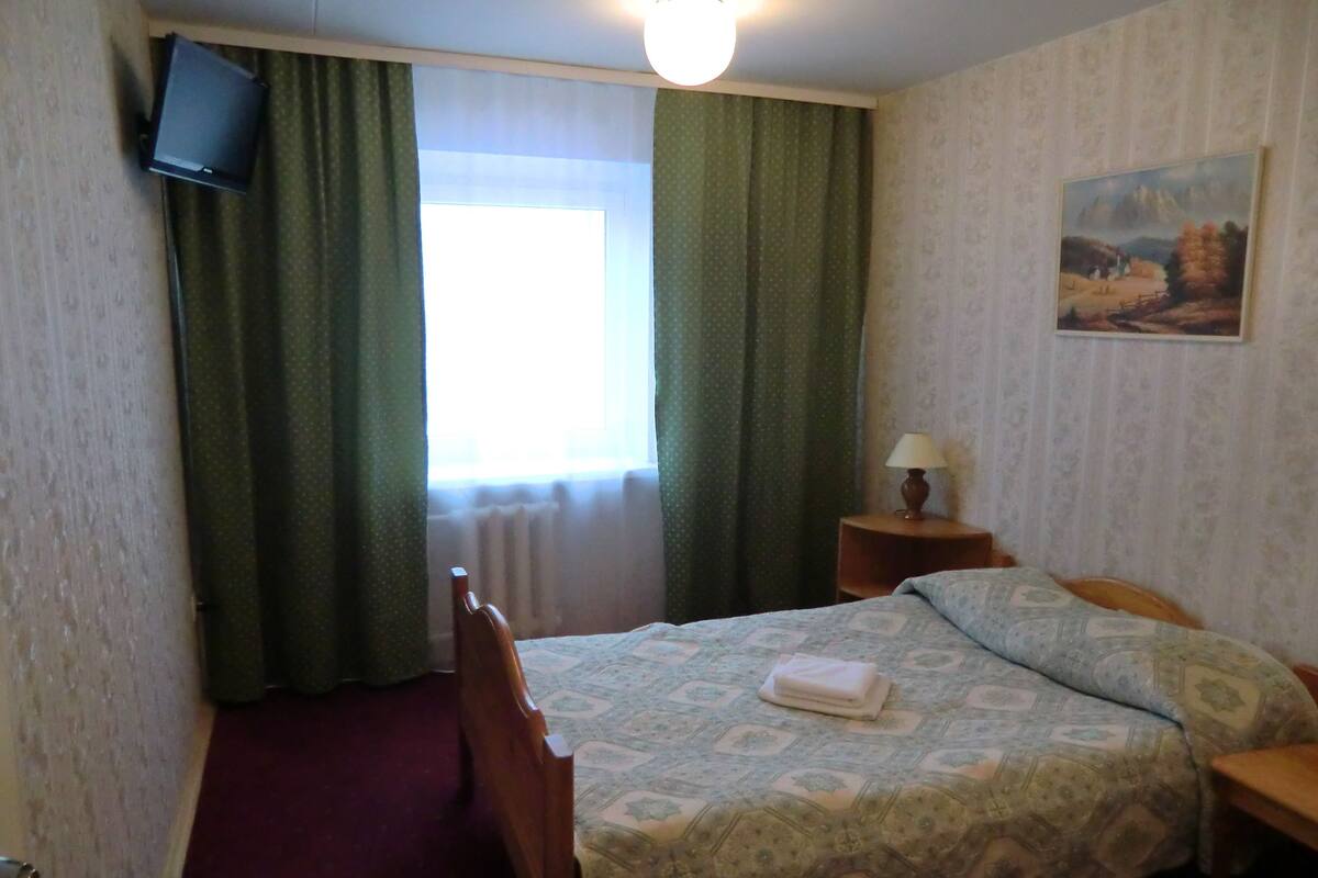 Ein Hotelzimmer mit Bett, Nachttisch, Lampe, einem Gemälde an der Wand und einem an der Wand befestigten Fernseher.