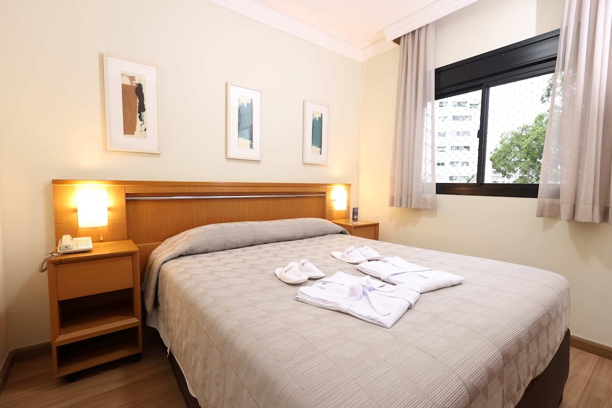 Ein Hotelzimmer mit einem Bett, einem Nachttisch mit Lampe, einem Fenster mit Vorhängen und einem Beistelltisch.