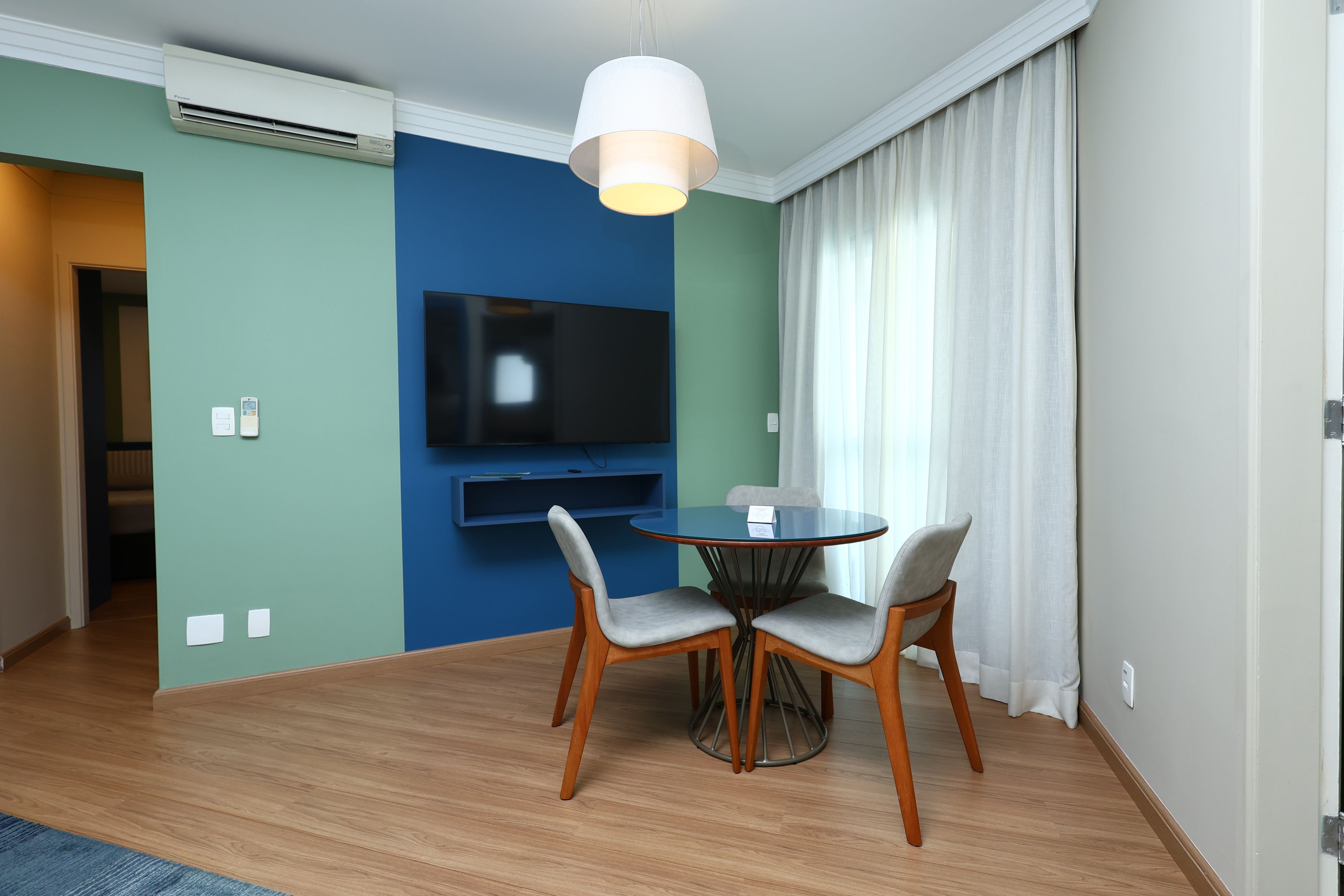 A modern hotel room with a dining area, a flat-screen TV mounted on the wall, and a large window with curtains.