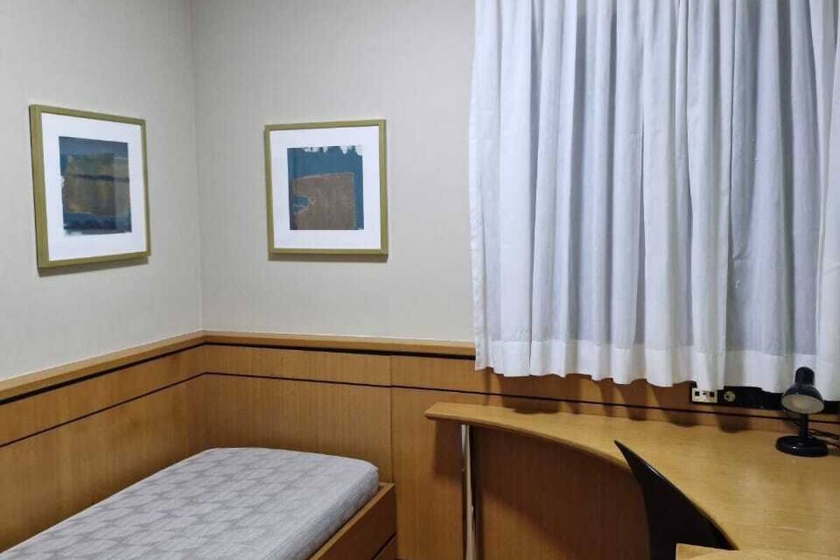 Ein kleines Zimmer mit einem Bett, einem Schreibtisch, einem Stuhl, einer Lampe und zwei gerahmten Bildern an der Wand.