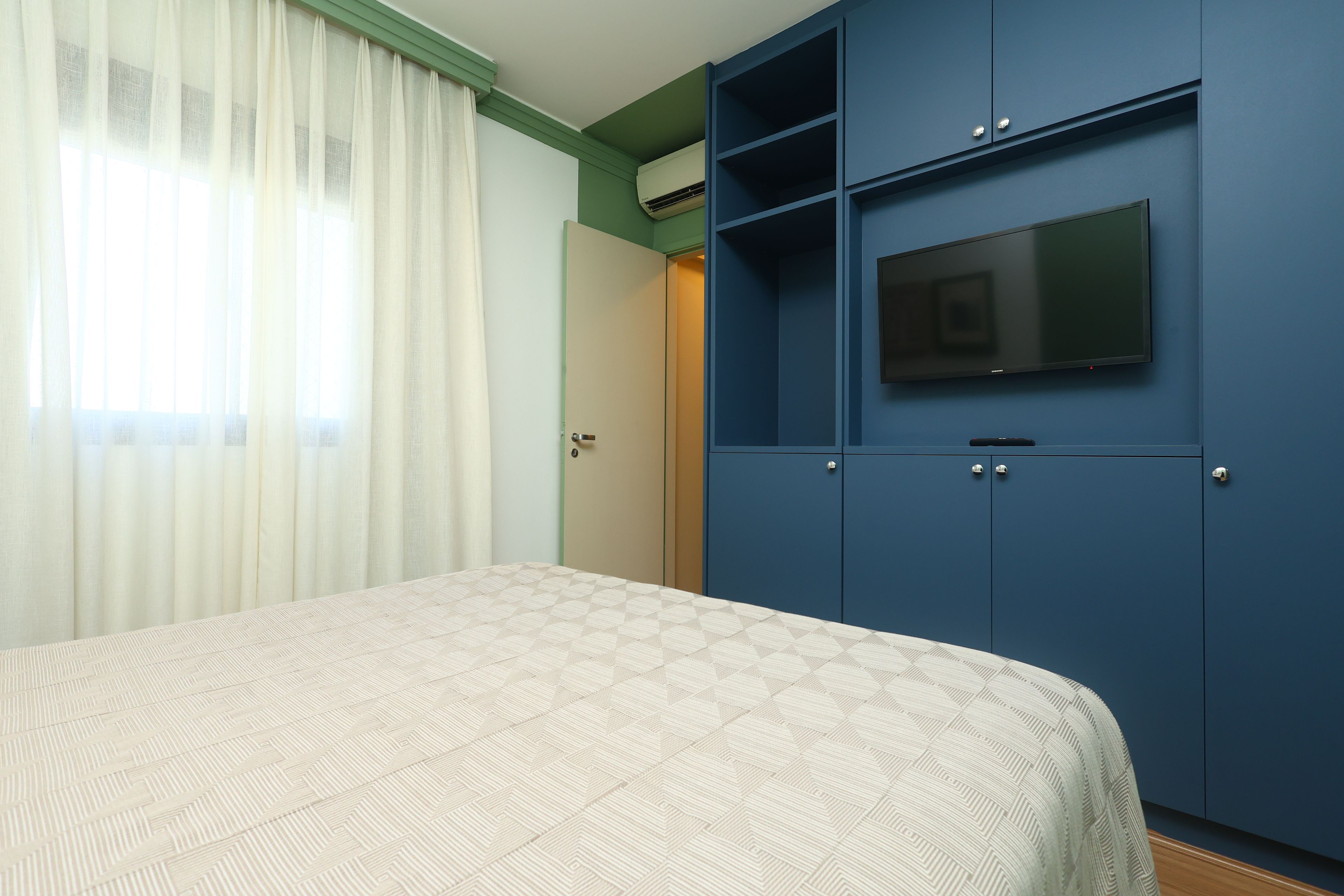 Un dormitorio con una cama, un televisor sobre un mueble azul y una ventana con cortinas traslúcidas.