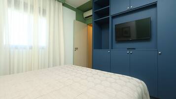 Un dormitorio con una cama, un televisor sobre un mueble azul y una ventana con cortinas traslúcidas.