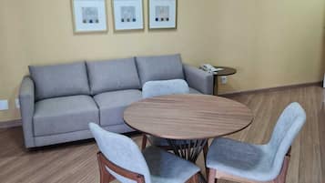 Una sala de estar con un sofá gris, una mesa redonda de madera y dos sillas grises. Hay tres cuadros enmarcados en la pared.