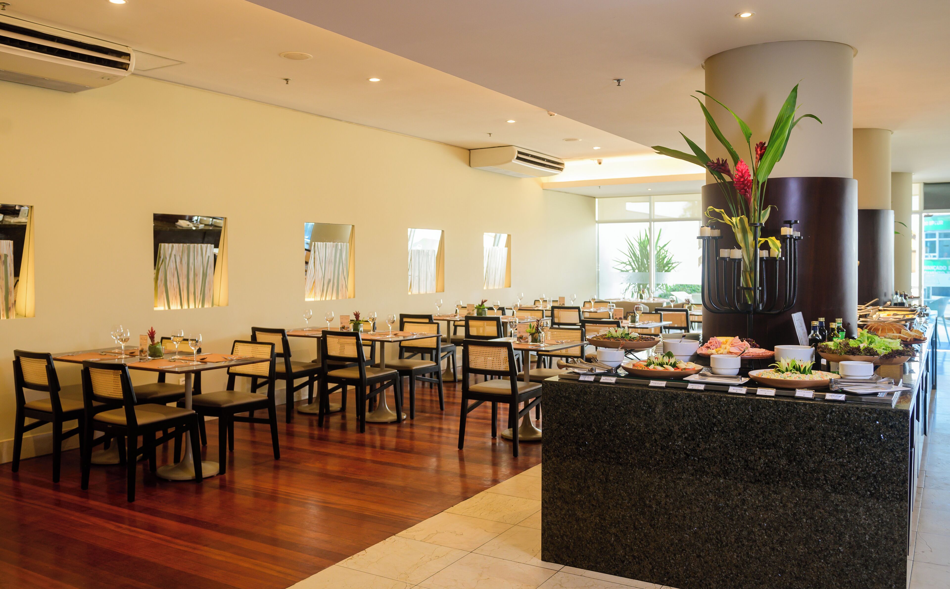 Un restaurante con varias mesas y sillas, una zona central de servicio con comida y bebidas, y amplios ventanales que permiten la entrada de luz natural.