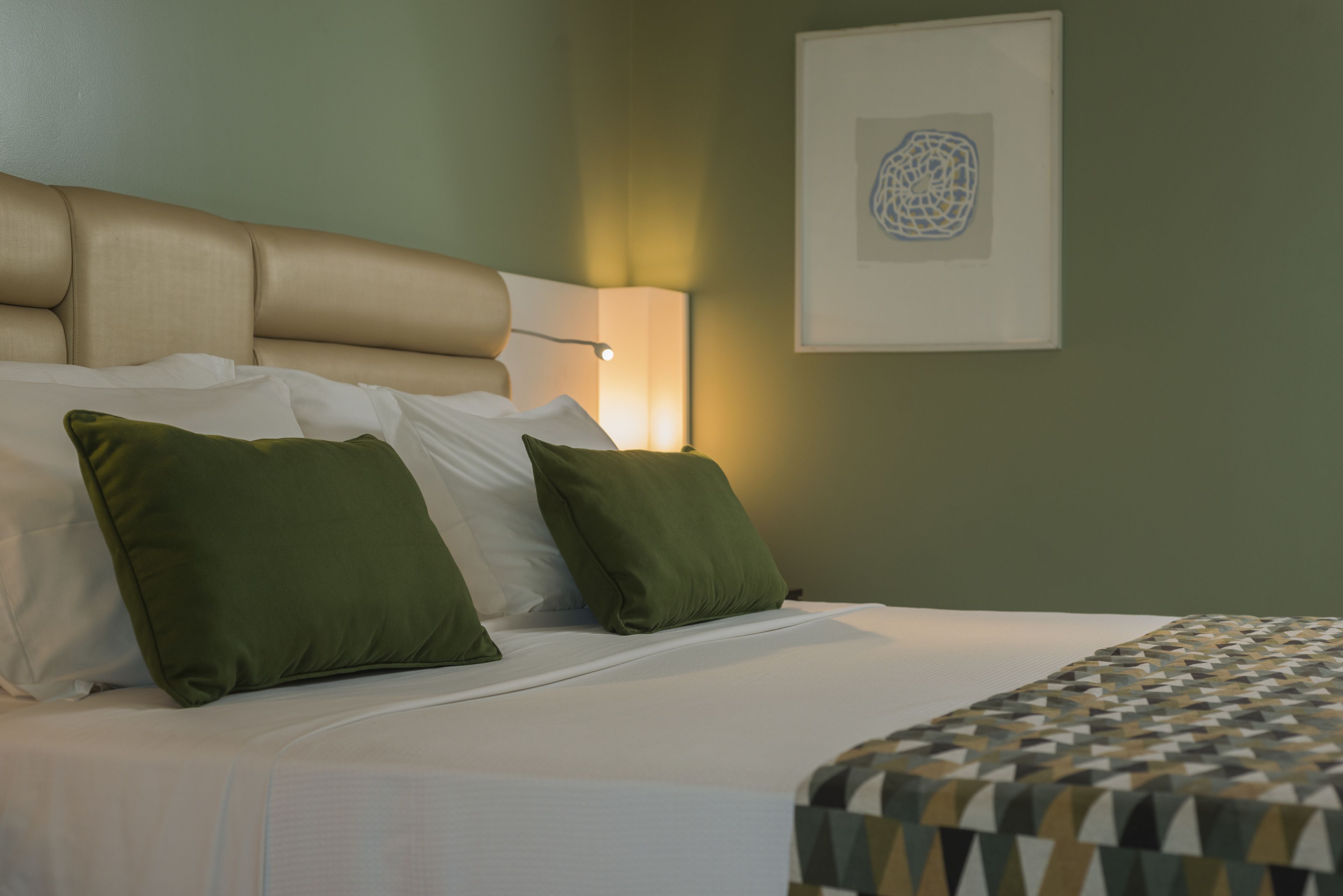 Een opgemaakt bed met witte en groene kussens, een bedsprei met dessin, en een hoofdbord.