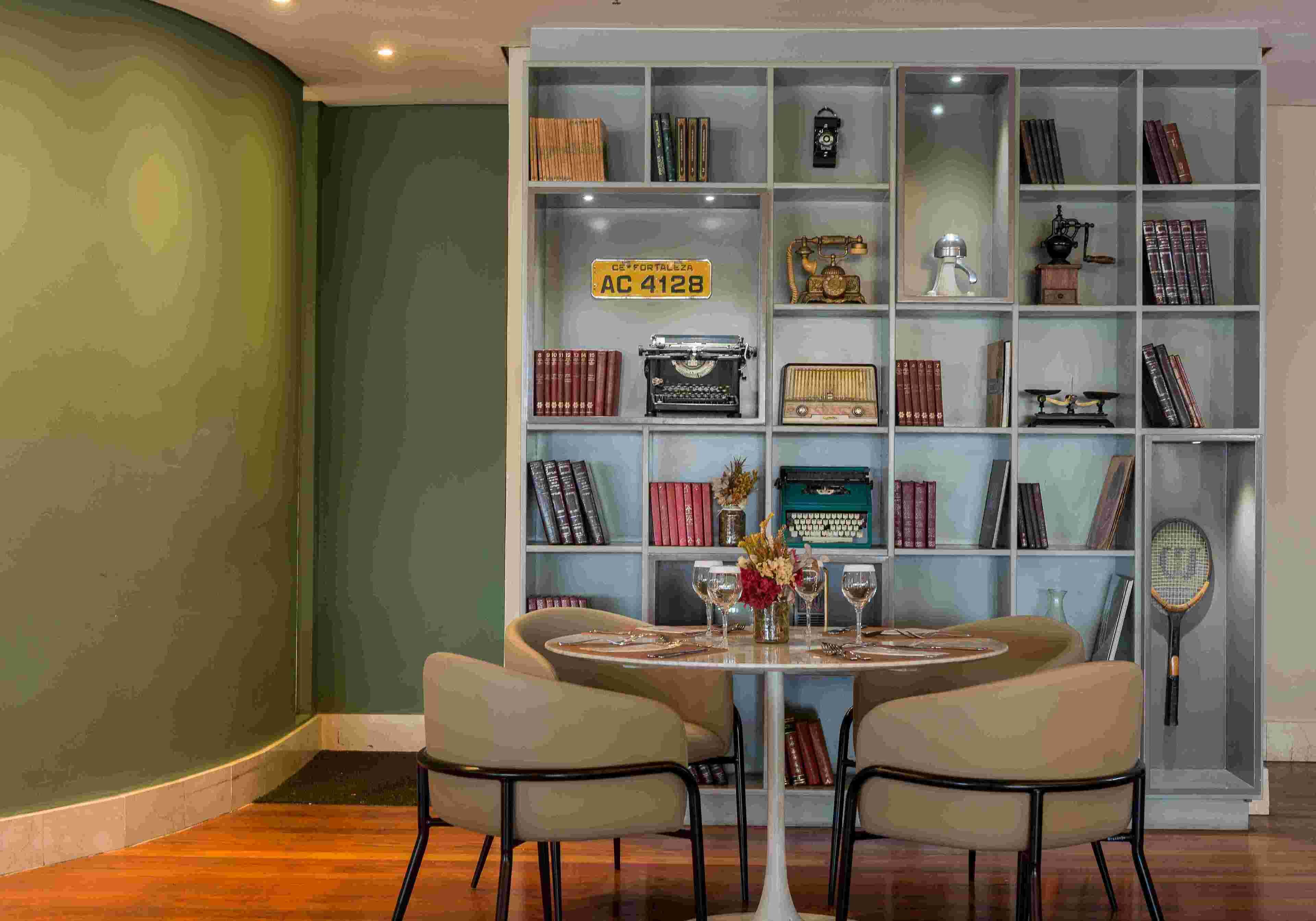 Una habitación con una mesa redonda, dos sillas, una estantería llena de libros y objetos antiguos, y una raqueta de tenis en la pared.