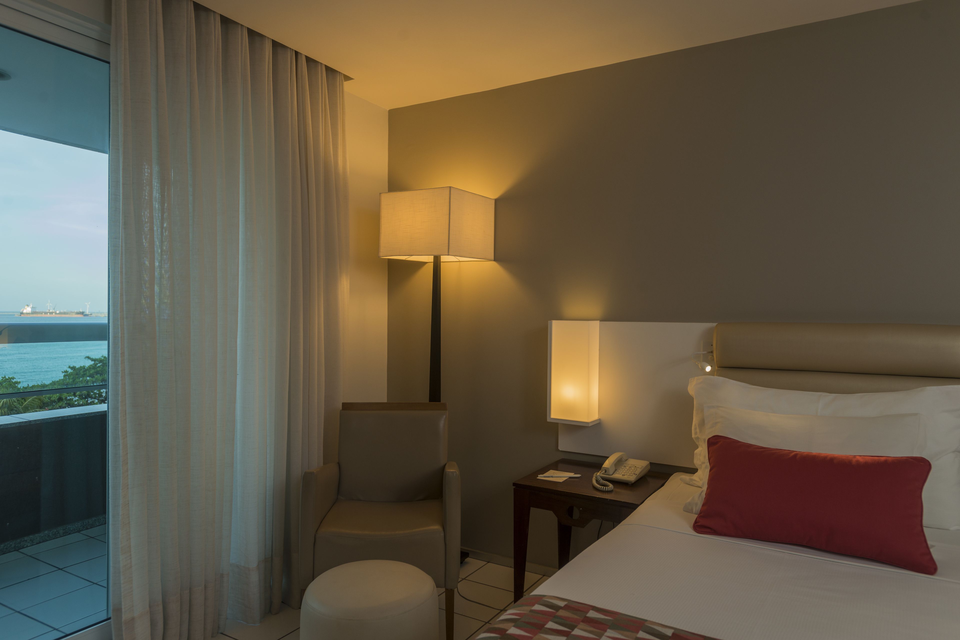 Habitación de hotel con cama, una silla, mesita de noche, lámpara y vista al mar.