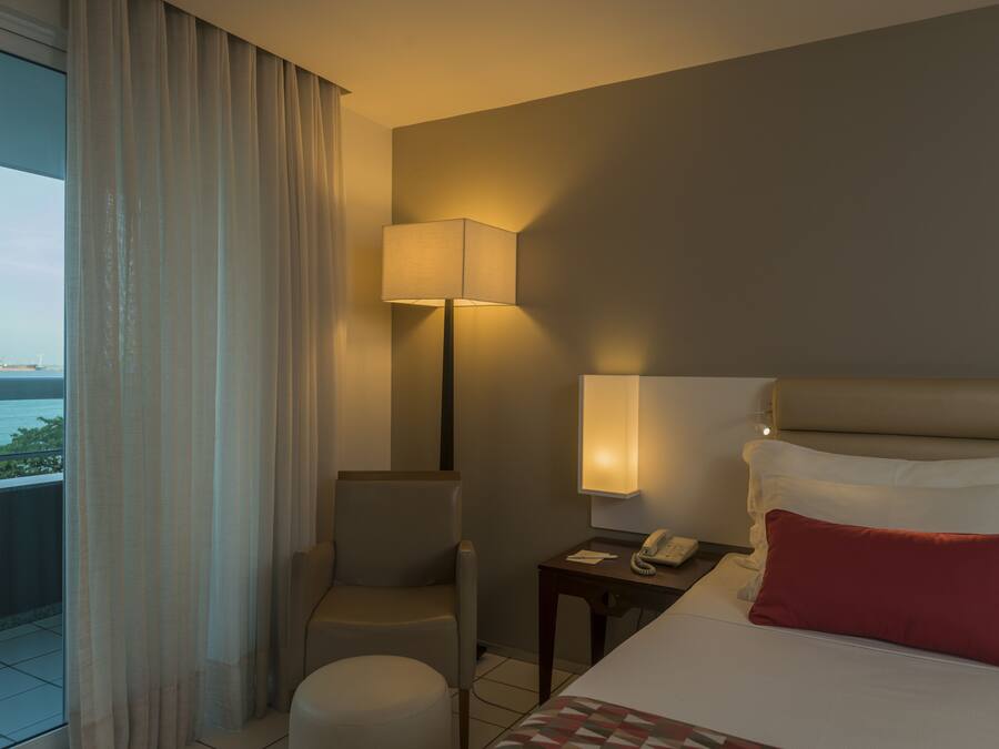 Habitación de hotel con cama, una silla, mesita de noche, lámpara y vista al mar.