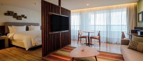 Una habitación de hotel con cama, televisión, zona de comedor con sillas, sofá y vistas al exterior a través de amplias ventanas.