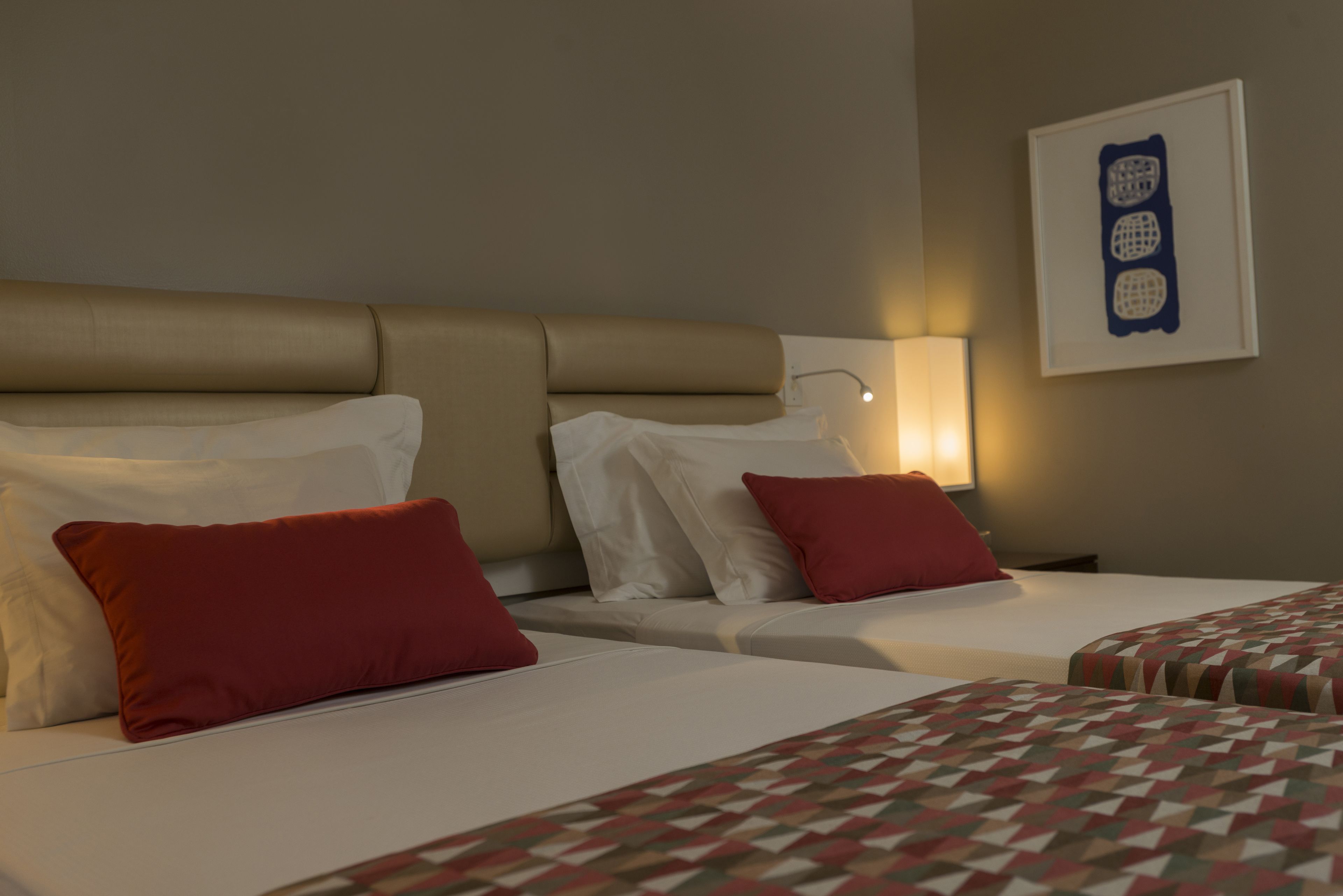 A hotel room with two beds, each with white linens and red pillows. There is a wall-mounted light fixture and a framed artwork above the beds.