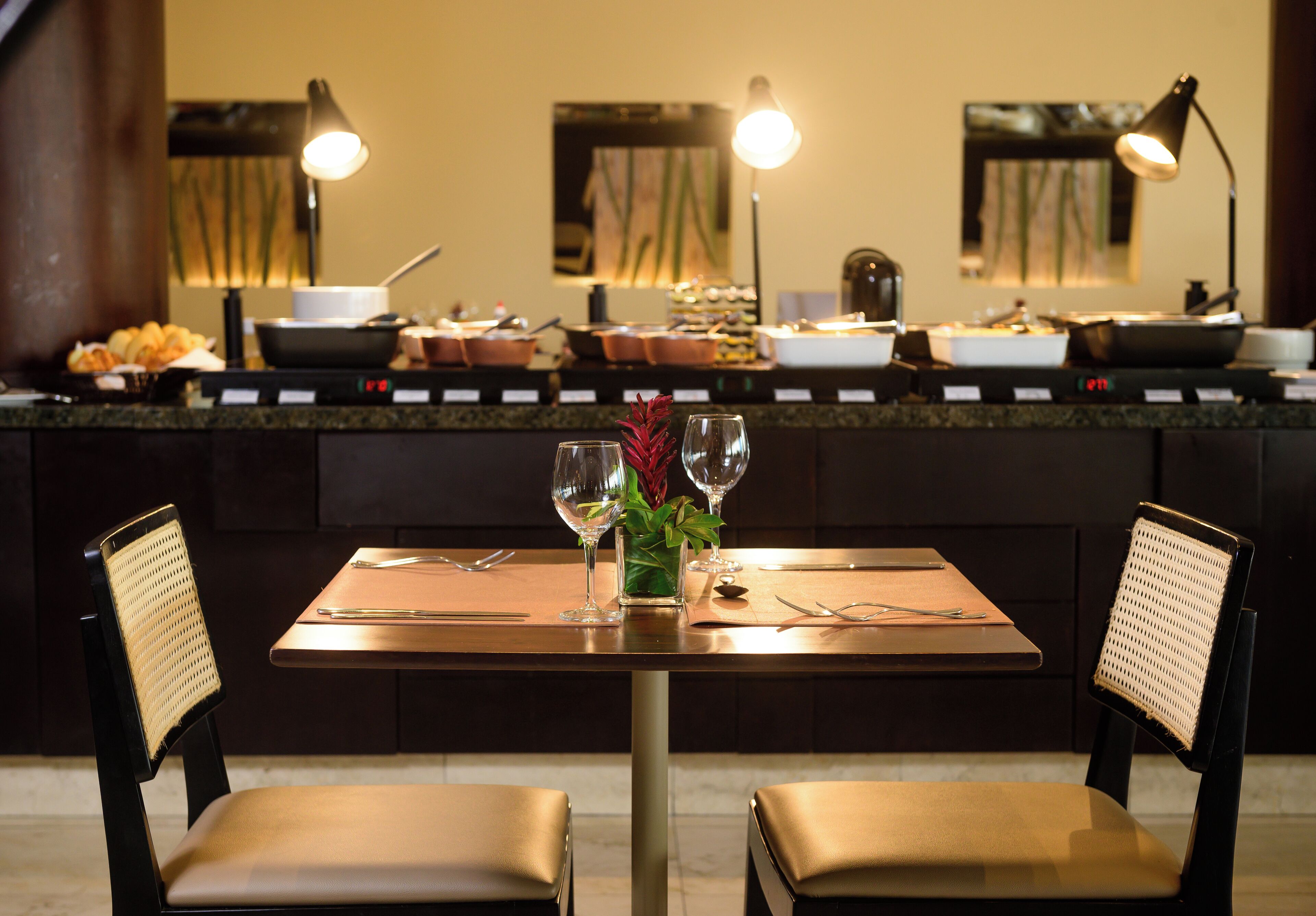 A dining area with a table set for two, two chairs, a vase with flowers, and a variety of dishes on the counter.
