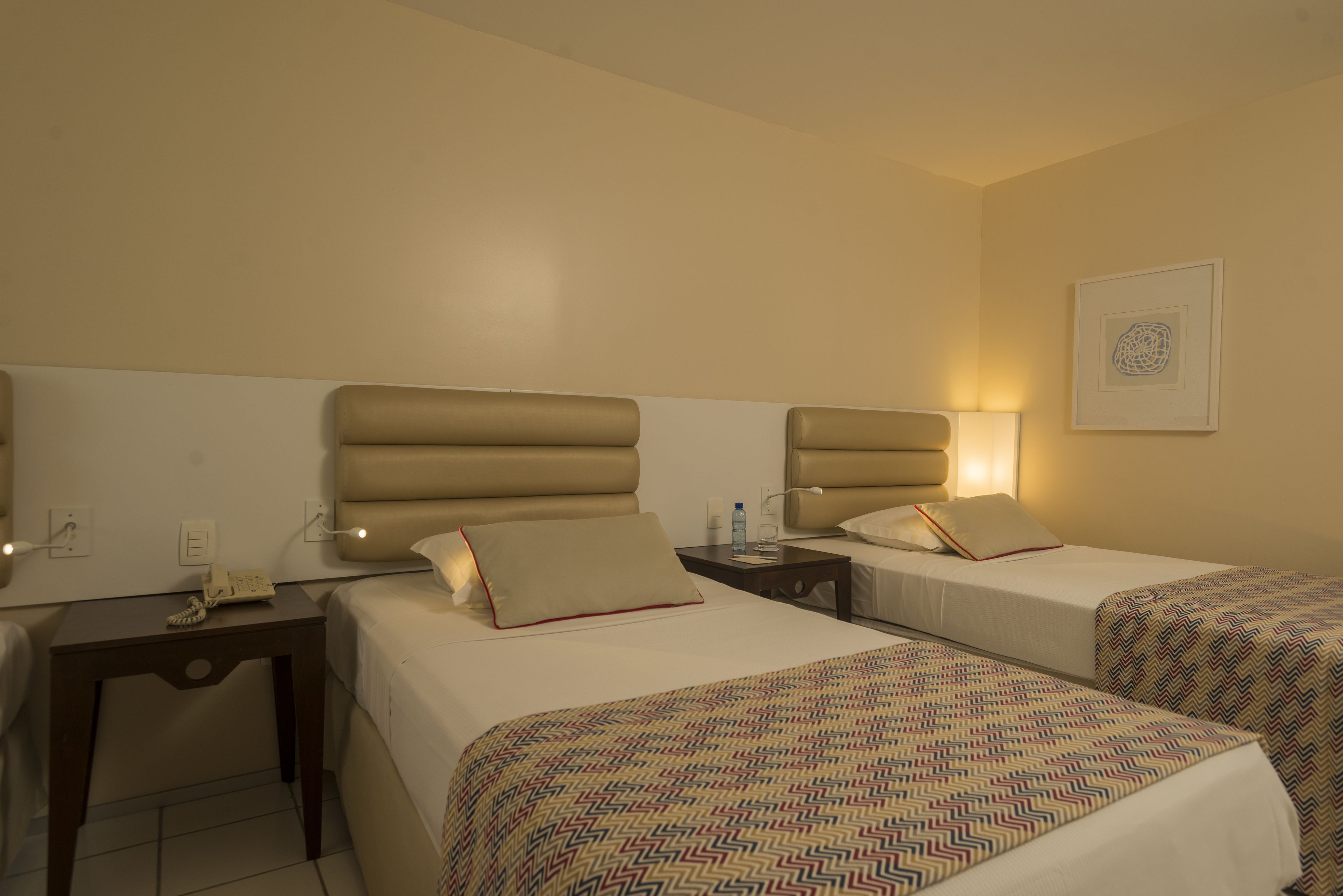 A hotel room with two beds, a bedside table with a lamp, and a wall-mounted artwork.