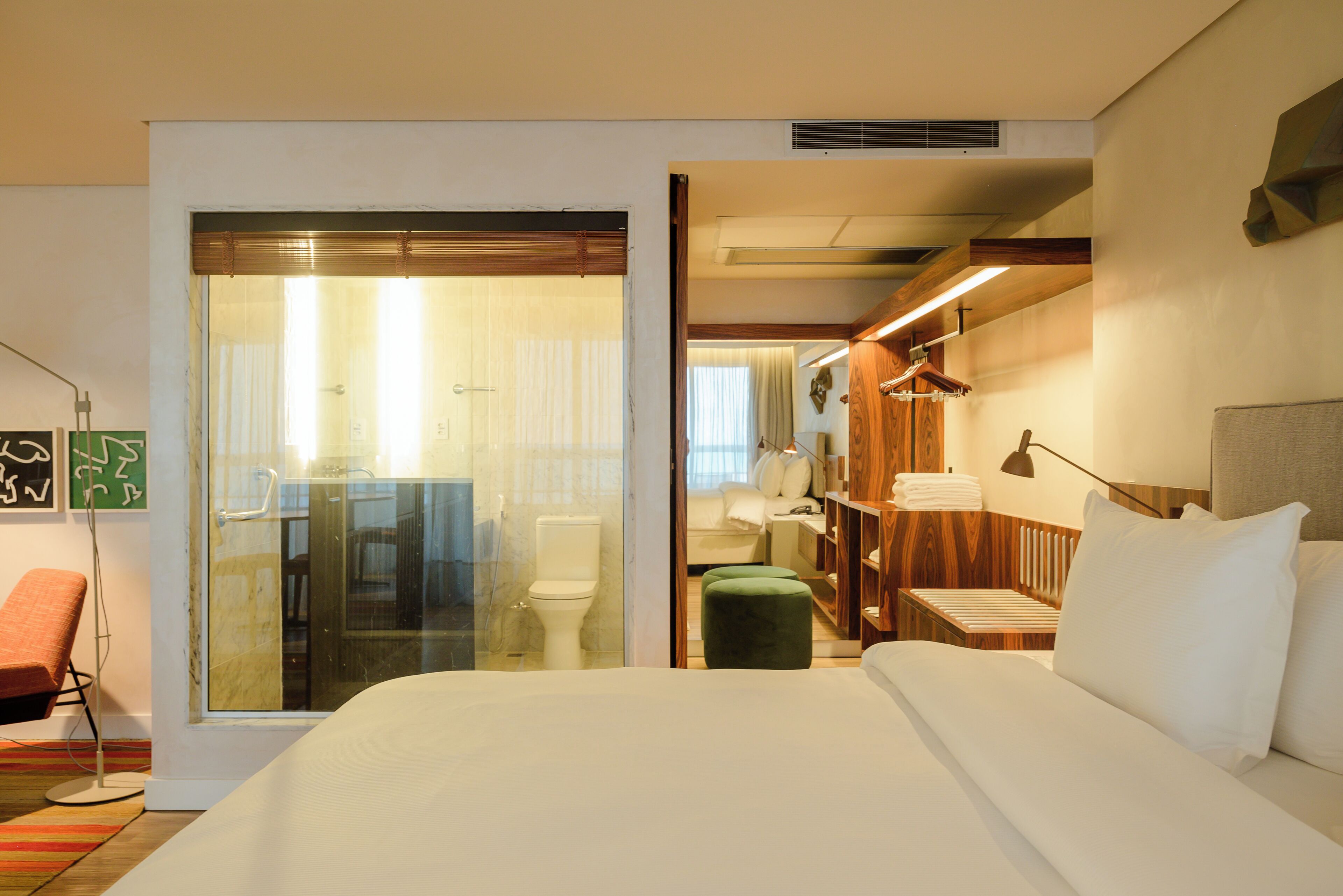 A hotel room with a large bed, a green ottoman, a wooden nightstand, and a glass shower enclosure.