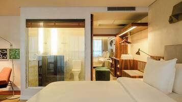 A hotel room with a large bed, a green ottoman, a wooden nightstand, and a glass shower enclosure.