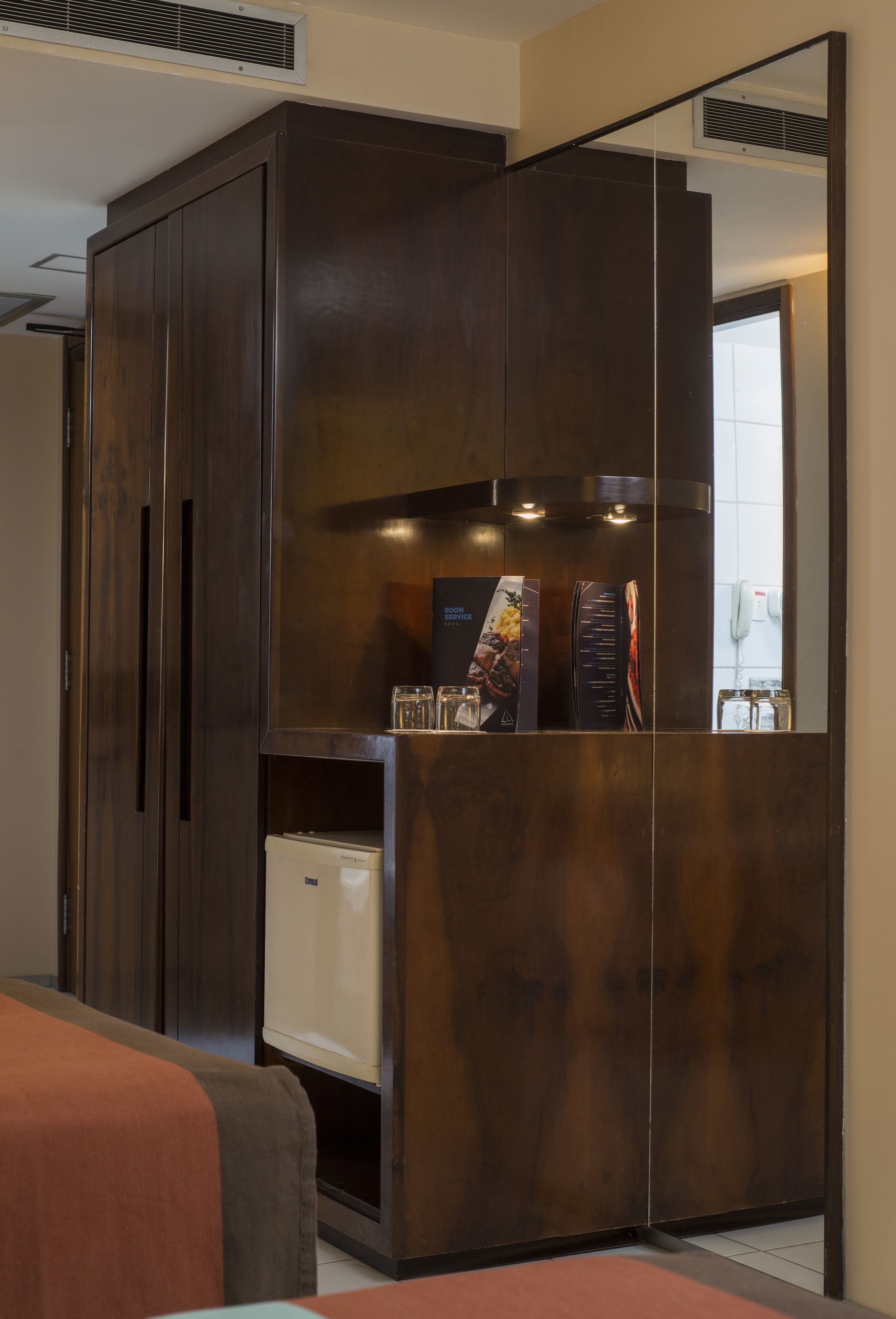 A compact room with a dark wooden wardrobe, a small white appliance, and a mirror reflecting a window and a phone.