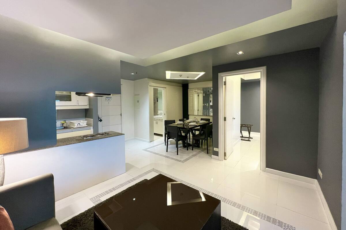 A modern living room with a black coffee table, a dining area with a white table and chairs, and a kitchen visible through an open door.