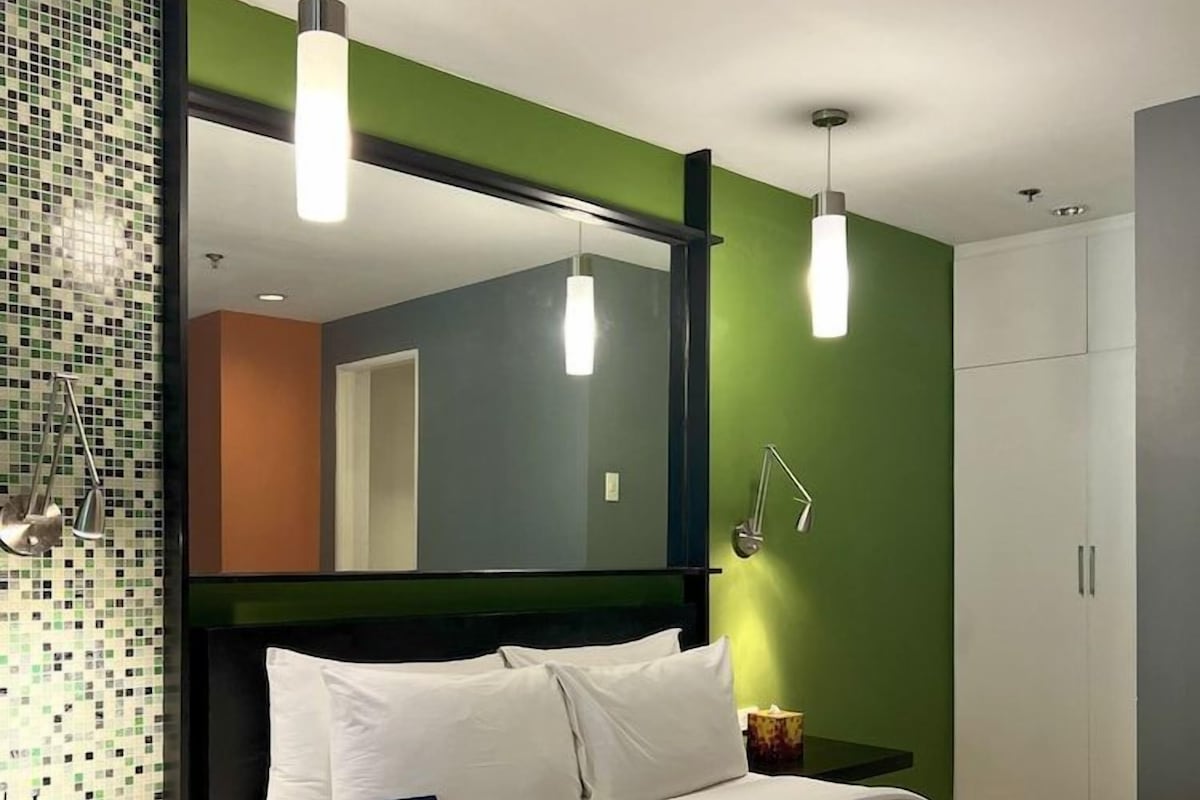 Una habitación de hotel moderna con una cama grande, mesita de noche, lámpara y un espejo.