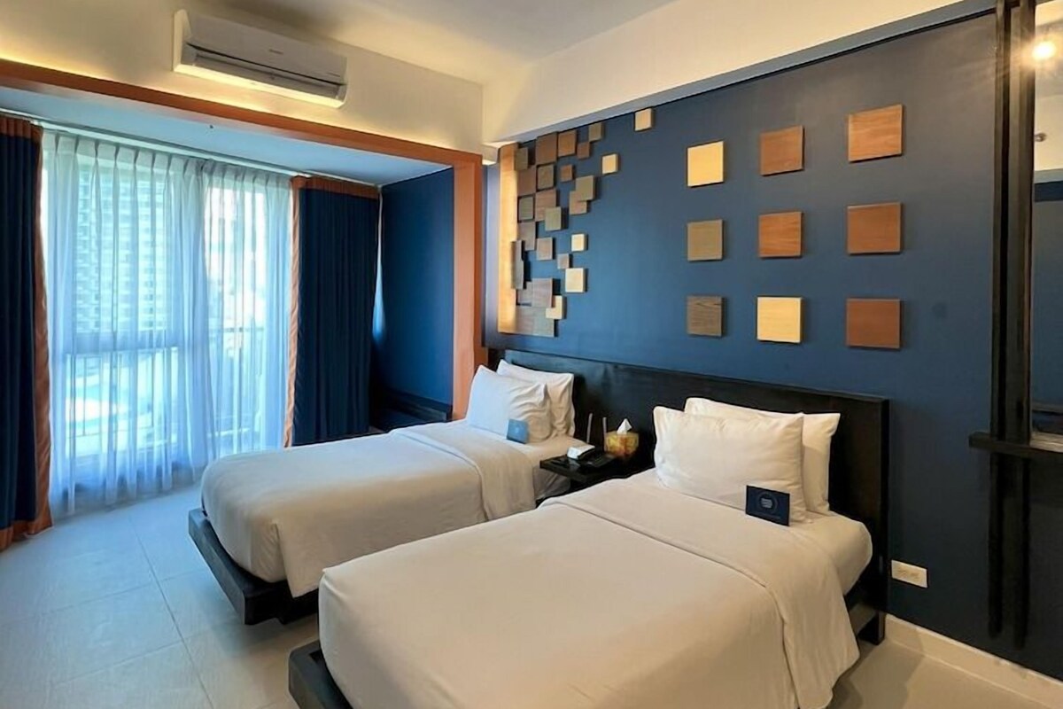 A hotel room with two beds, a large window with curtains, a wall-mounted air conditioner, and a modern light fixture.