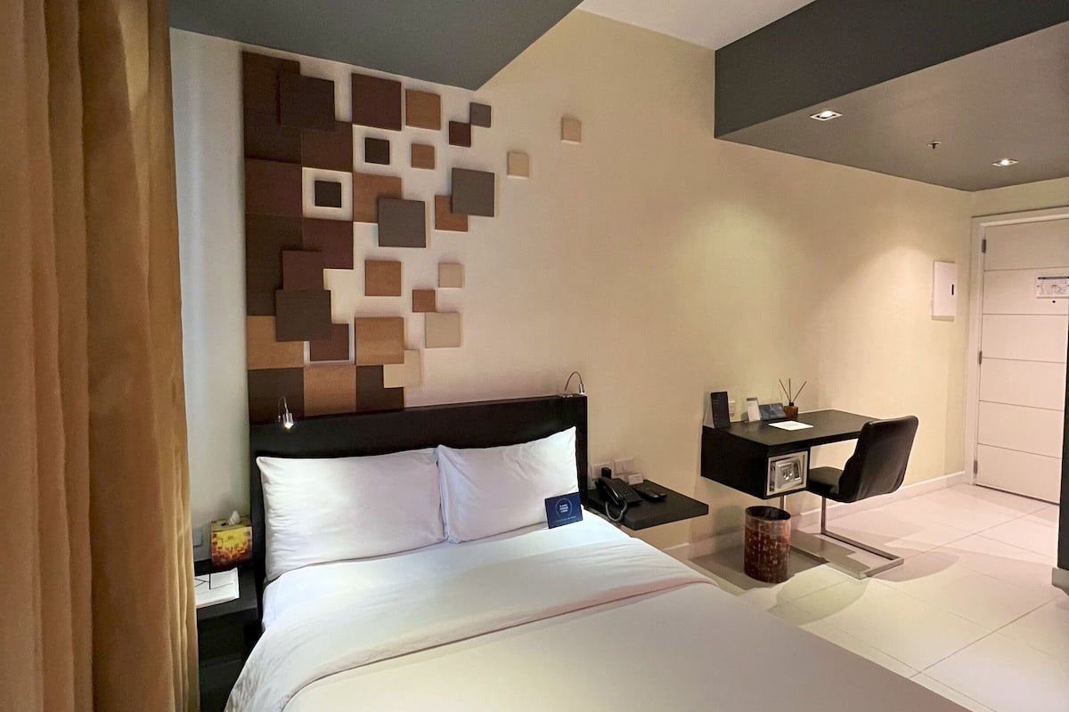A modern hotel room with a large bed, a desk with a chair, and a wall-mounted light fixture.
