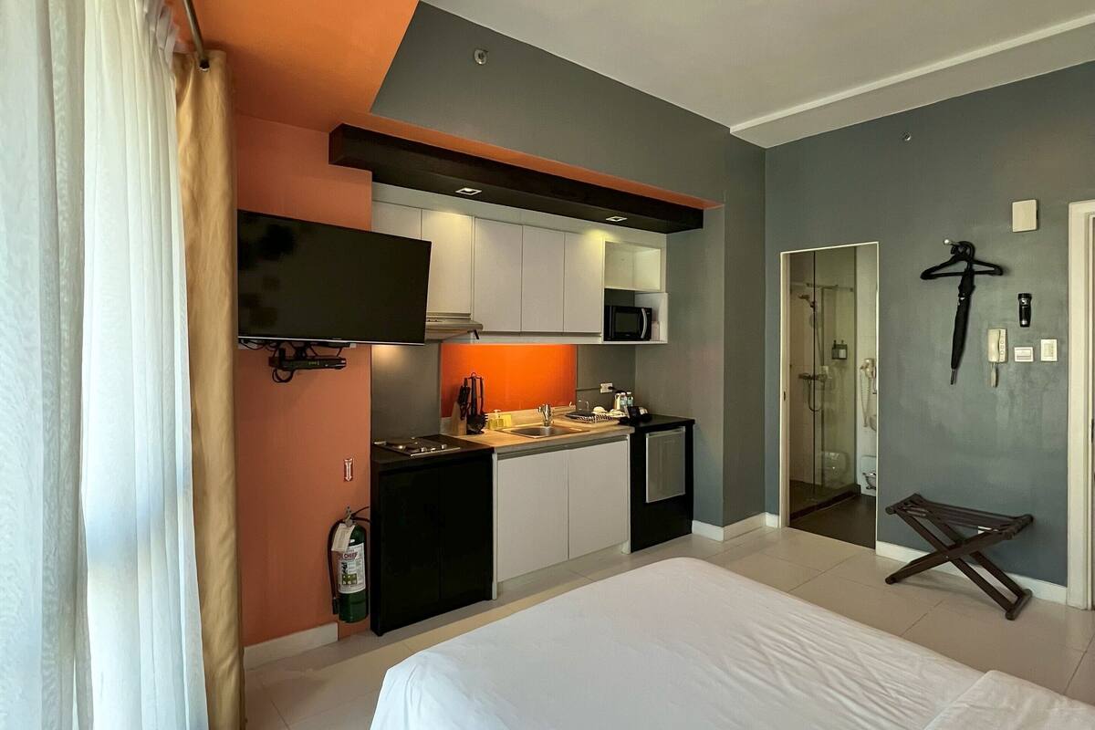 A modern hotel room with a large bed, a kitchenette, and a shower area.