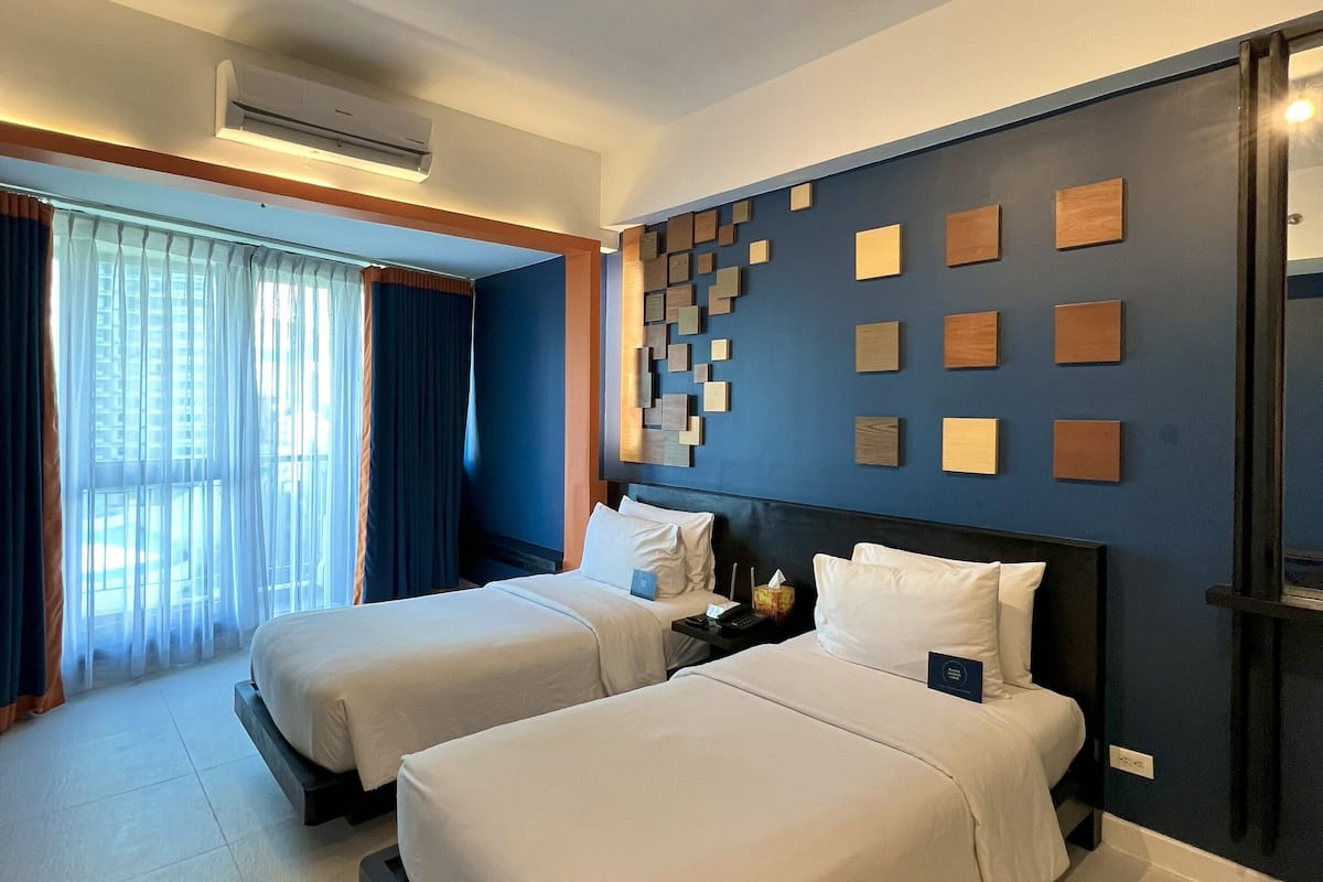 A hotel room with two beds, a large window with curtains, a wall-mounted air conditioner, and a series of framed pictures.