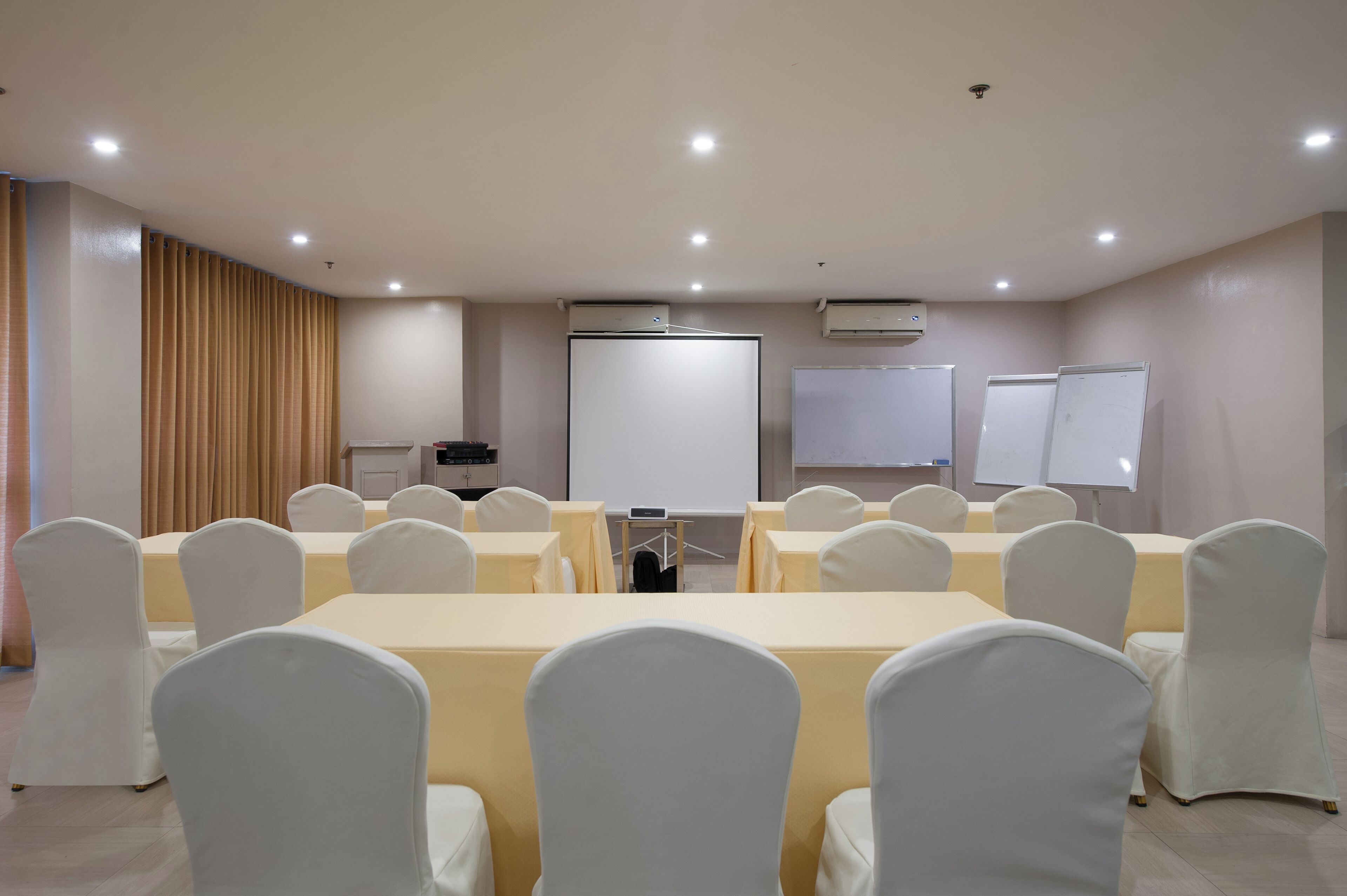 Un salón de conferencias con una larga mesa, sillas blancas, una pantalla de proyector y pizarras.