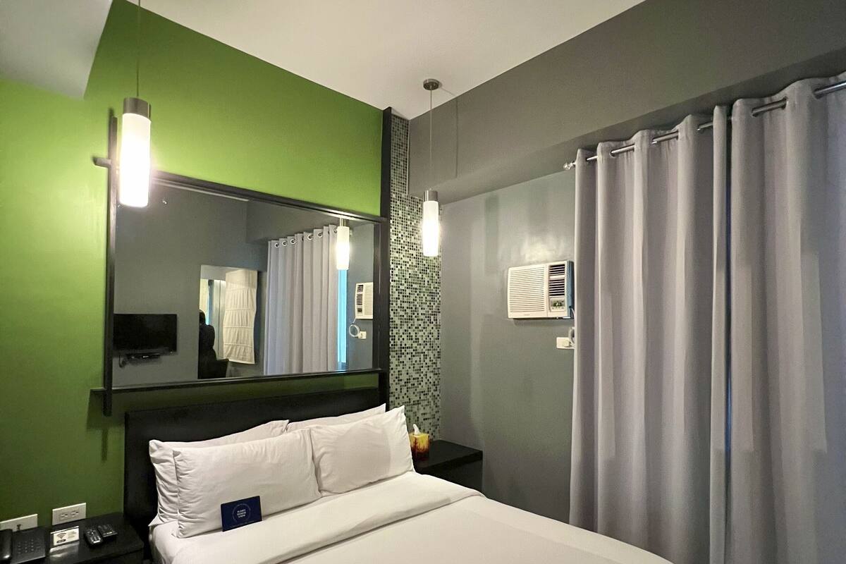 A modern hotel room with a large bed, green walls, a mirror, a flat-screen TV, and a window with curtains.
