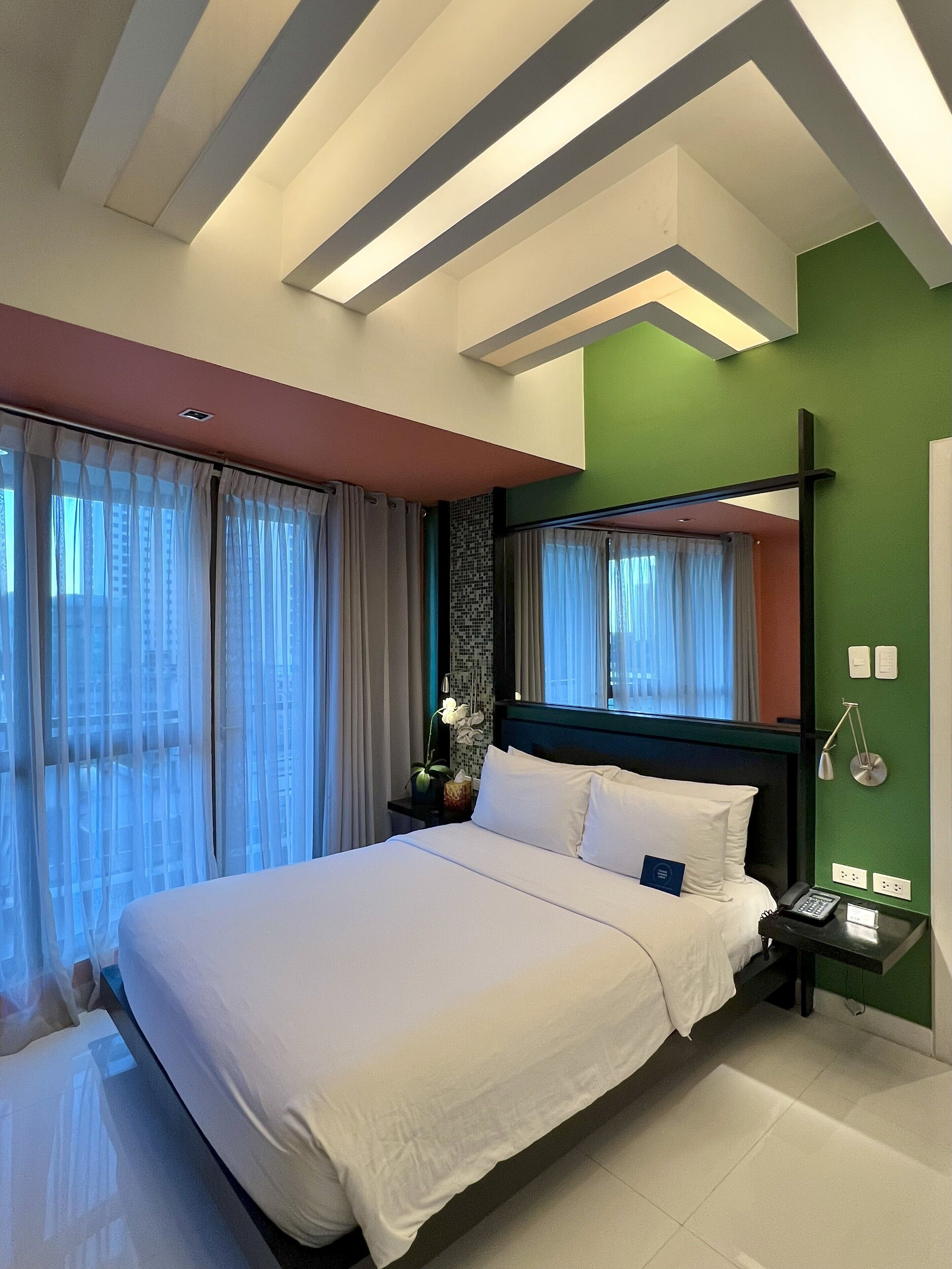 Une chambre d’hôtel moderne avec un grand lit, une table de chevet avec un téléphone et un mur d’accent vert.
