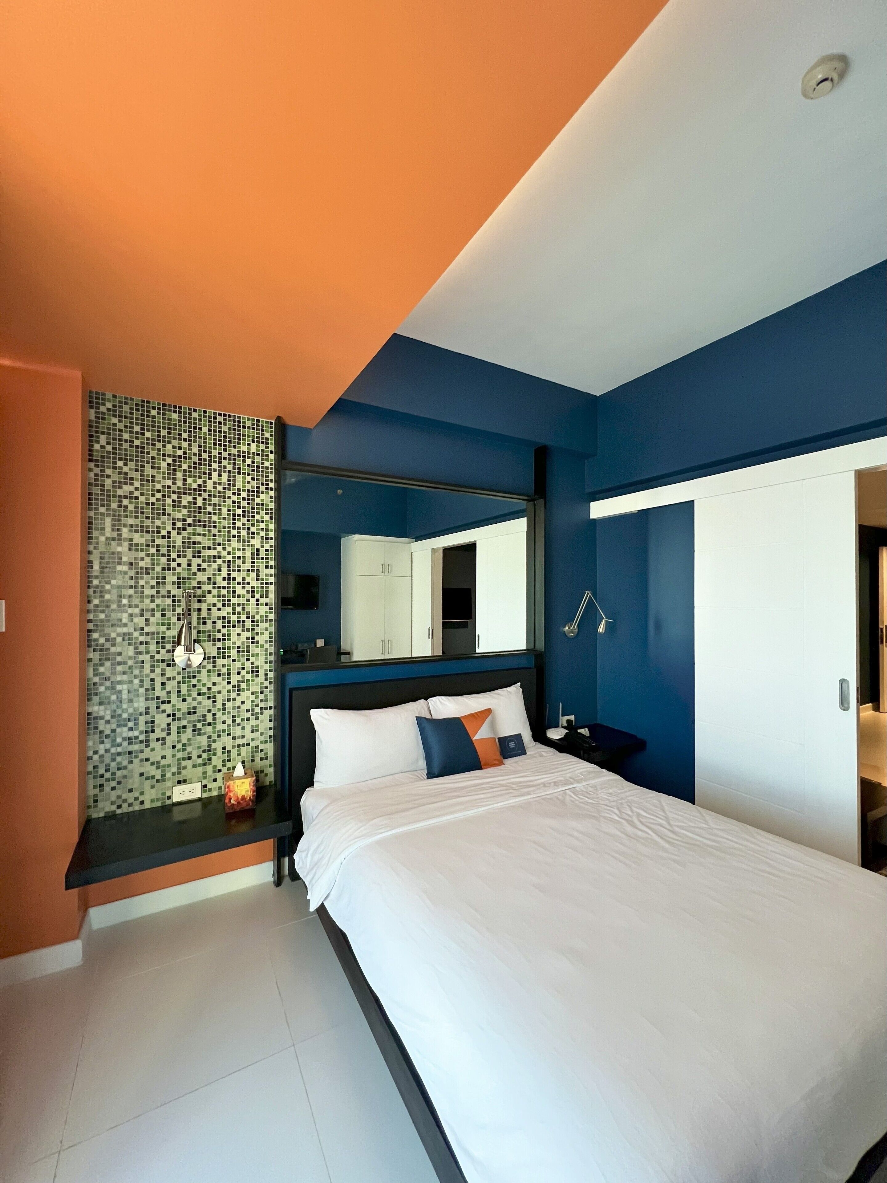 A modern hotel room with a large bed, a nightstand, a mirror, and a bathroom door.