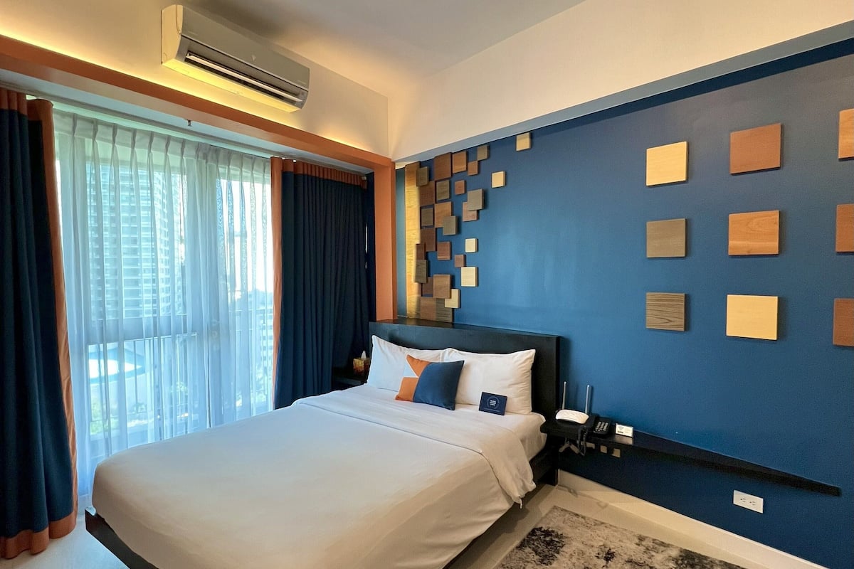 A modern hotel room with a large bed, a blue accent wall with wooden panels, a black and white rug, and a window with curtains.