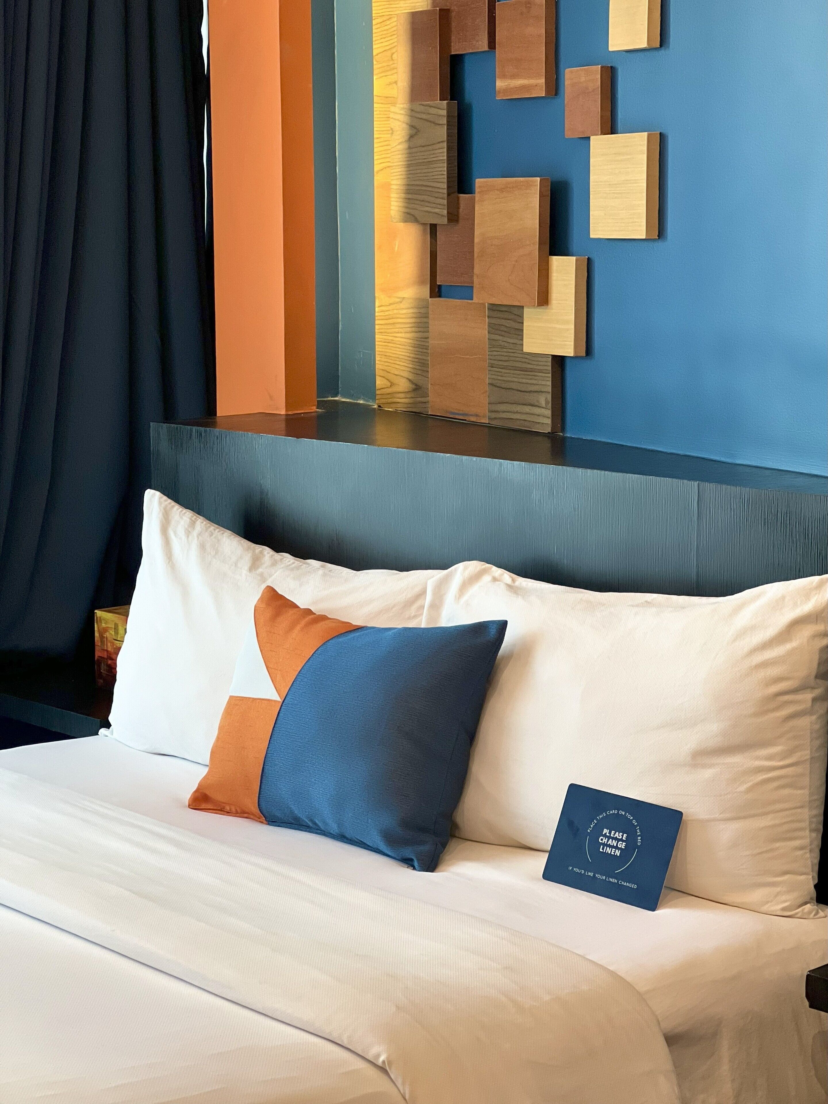 A neatly made bed with white and blue pillows, a blue card on the bed, and a wall with a geometric wooden art piece.