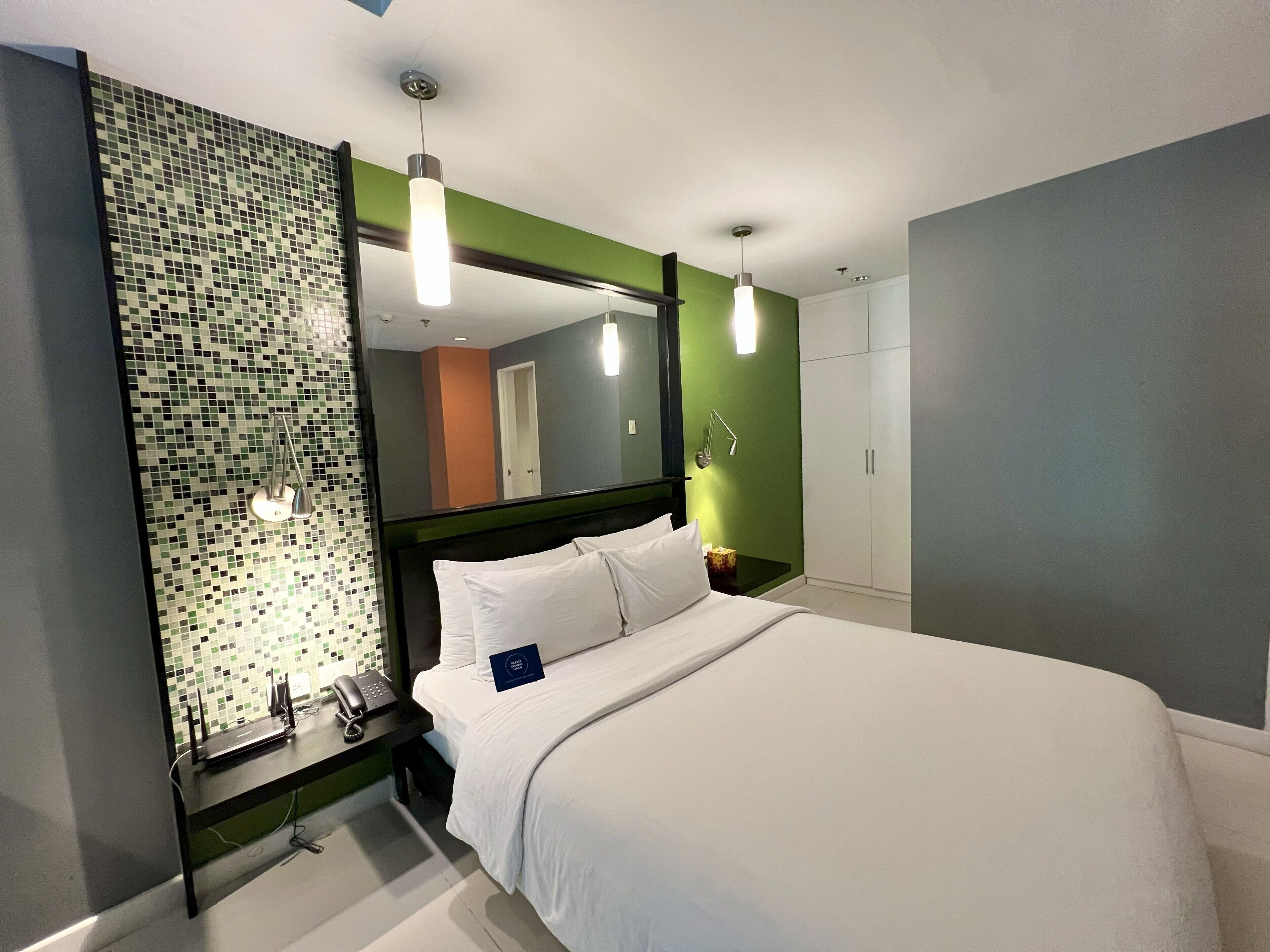 A modern hotel room with a large bed, a bedside table with a phone, a mirror, and a green accent wall.