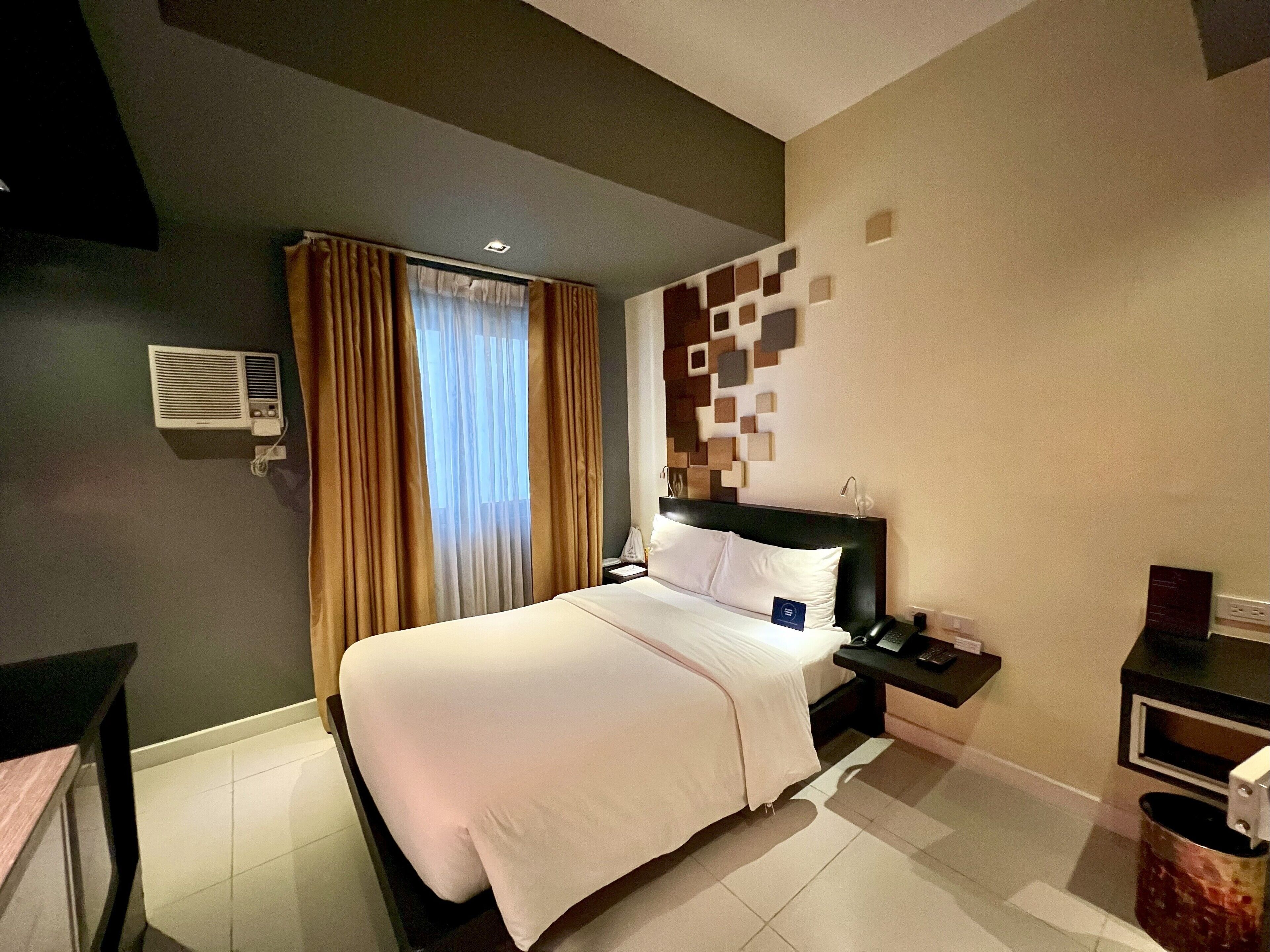 A hotel room with a bed, bedside table, and a wall-mounted air conditioning unit.
