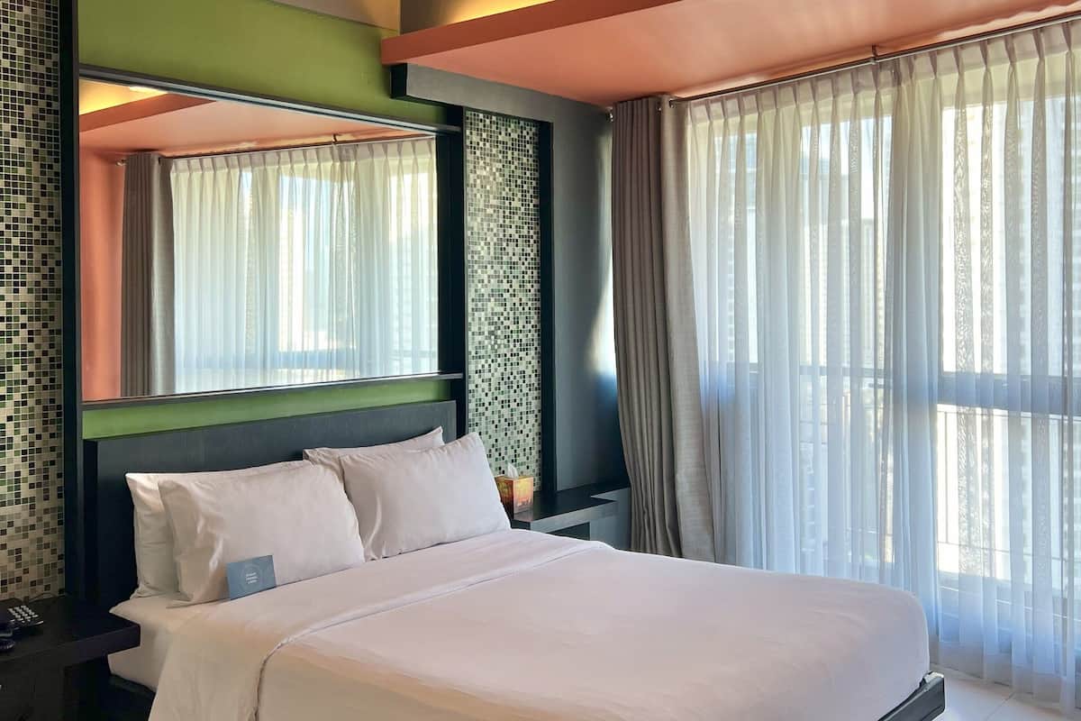 A hotel room with a large bed, an air conditioning unit, a nightstand, a mirror, and a window with curtains.