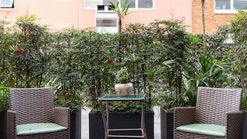 Uma área externa com mobiliário de vime e uma mesa de vidro, cercada por vegetação densa e um edifício ao fundo.