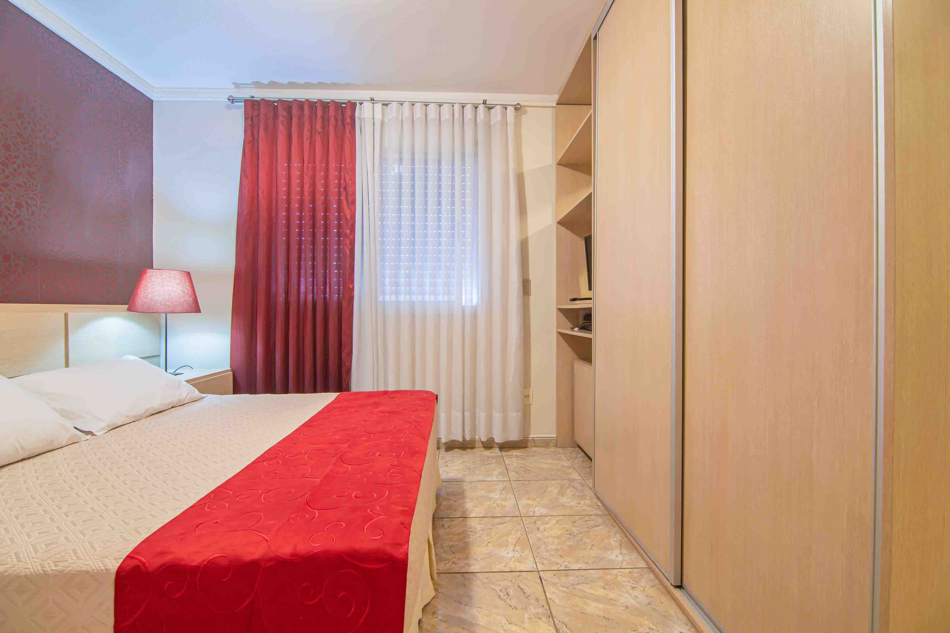 A hotel room with a bed, a nightstand, a lamp, a window with curtains, and a wardrobe.