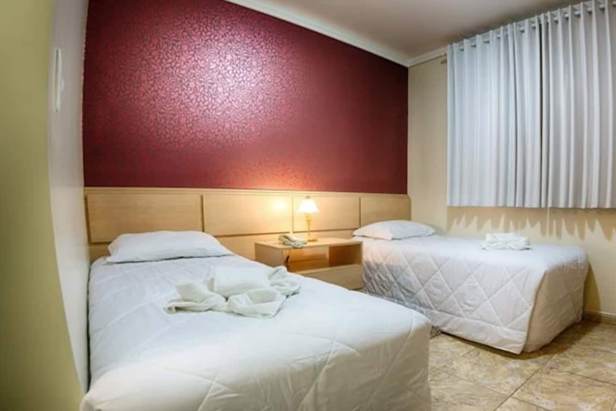 A hotel room with two beds, a nightstand with a lamp, a window with curtains, and a tiled floor.