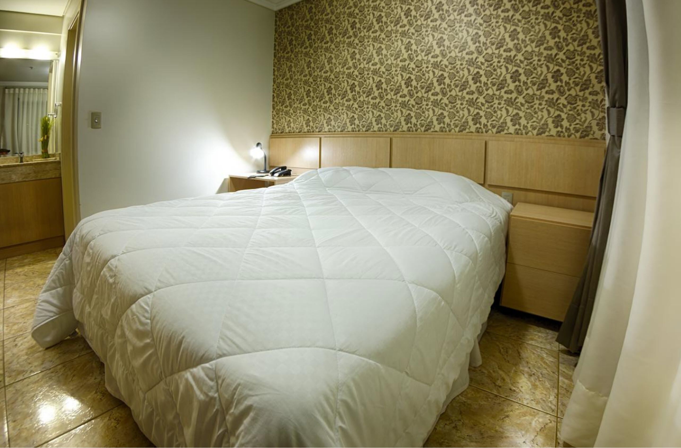 A hotel room with a large bed, a wooden headboard, a nightstand with a lamp, and a floral-patterned wall.
