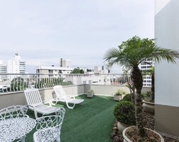 Una terraza en la azotea con mobiliario blanco para exteriores, una palmera y vistas a edificios de la ciudad.