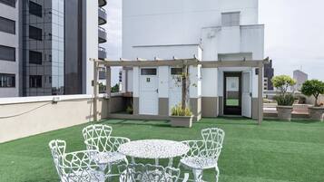 A rooftop garden with white metal chairs and tables, surrounded by a white building with balconies.