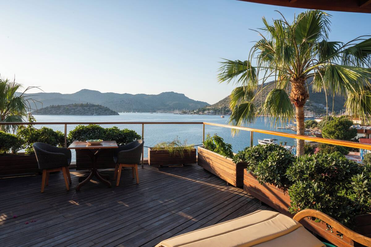 Eine Holzterrasse mit Tisch und Stühlen, umgeben von Pflanzenkübeln und einer Palme, mit Blick auf ein ruhiges Meer und ferne Hügel.