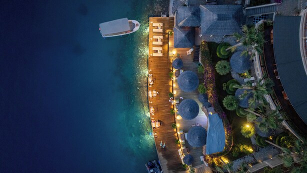 An aerial view of a waterfront hotel with a wooden deck, umbrellas, and a boat docked nearby.