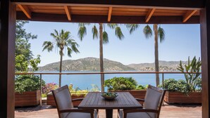 A wooden deck with a table and chairs, overlooking a body of water and hills.