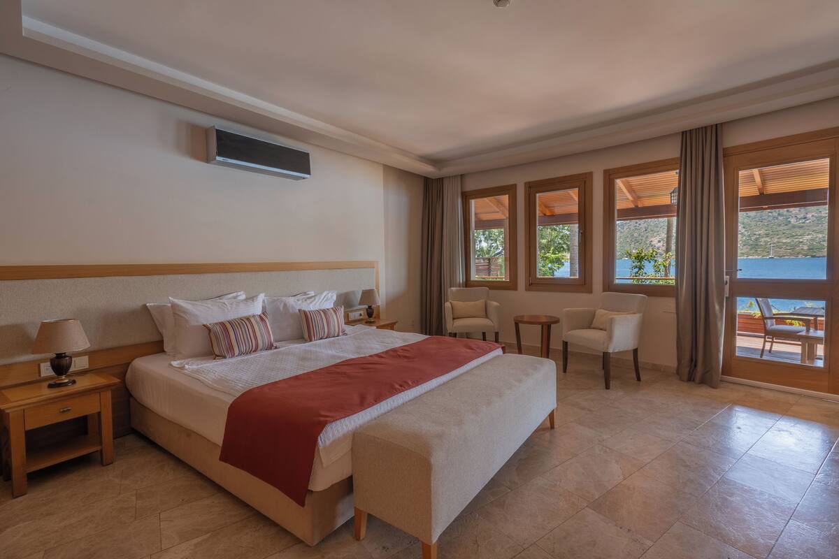 Ein Hotelzimmer mit einem großen Bett, einem Nachttisch mit Lampe, einem an der Wand montierten Fernseher und Blick ins Freie durch große Fenster.