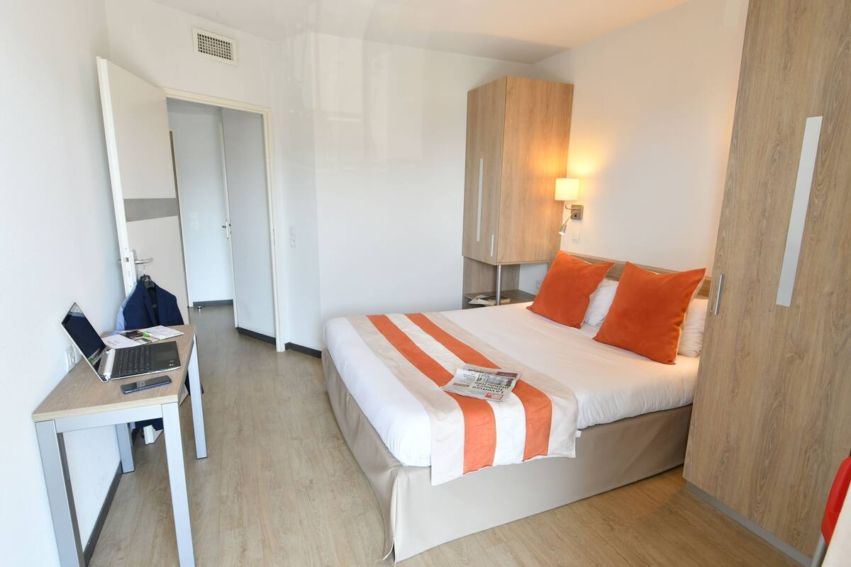 A hotel room with a bed, a desk with a laptop, a wardrobe, and a bedside lamp.