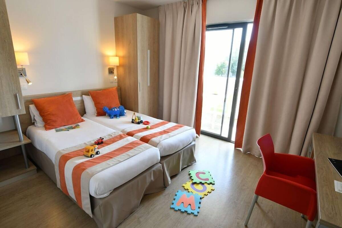 A hotel room with two beds, a desk, a chair, and alphabet foam mats on the floor.