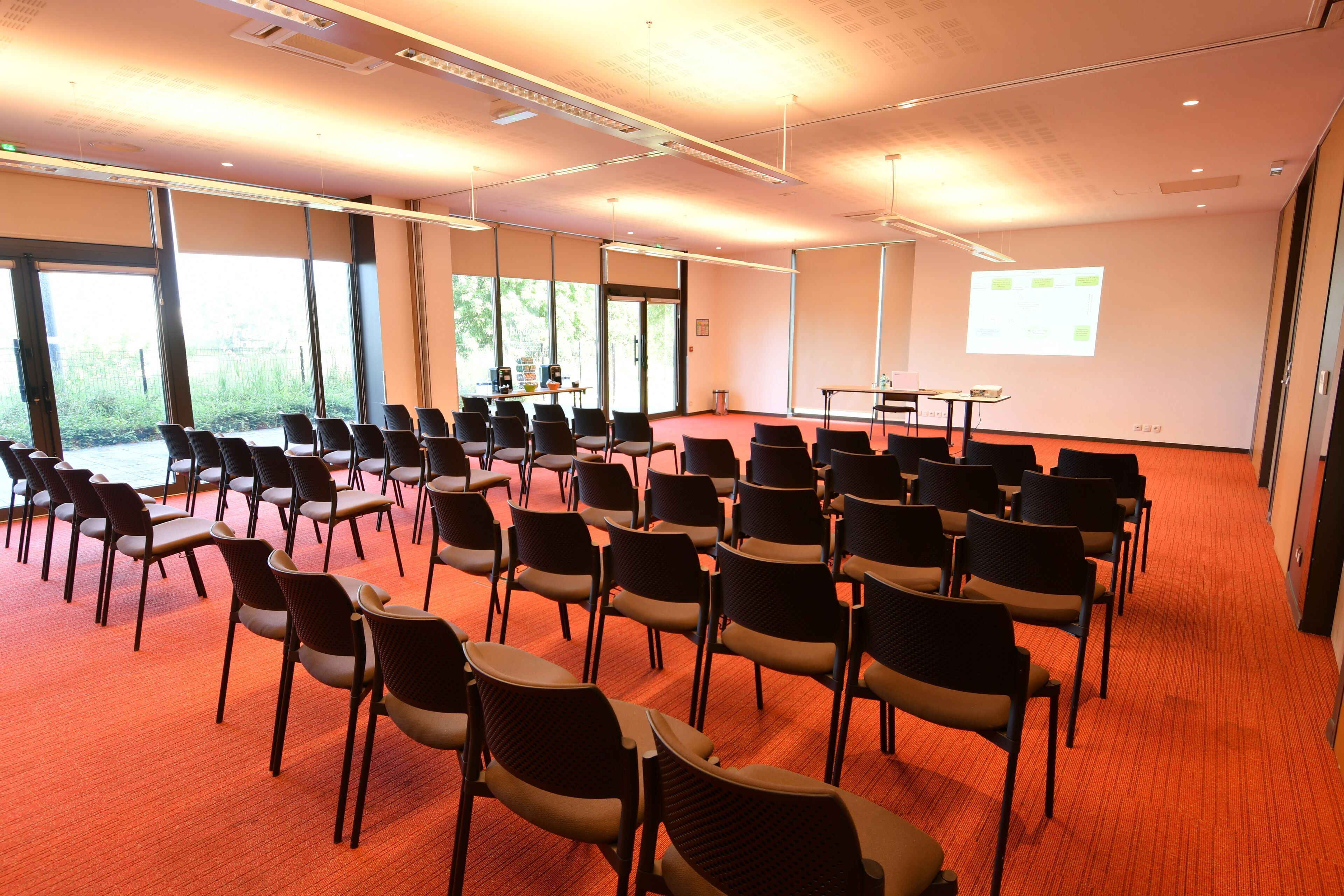 Une salle de conférence avec des rangées de chaises, un écran de projection et de grandes fenêtres.