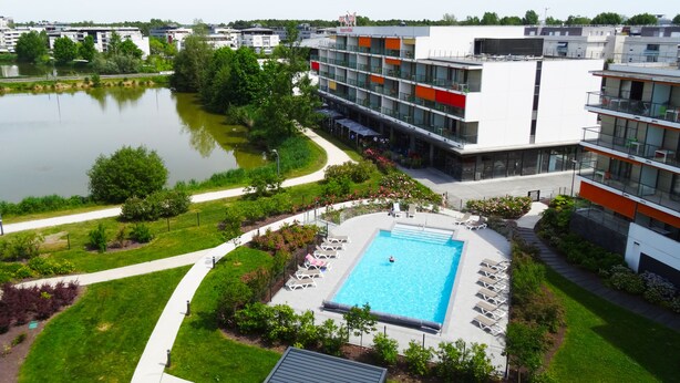 A modern hotel with a swimming pool, surrounded by greenery and a body of water. - Appart Hôtel Mer & Golf City Bordeaux Bruges (Bruges)