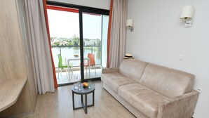 A room with a beige sofa, a small table with a fruit bowl, and a view of a body of water through a glass door. - Appart Hôtel Mer & Golf City Bordeaux Bruges (Bruges)