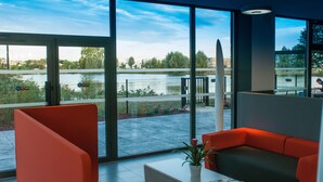 A modern interior with large windows, a seating area with orange and gray furniture, and a view of a body of water and greenery outside. - Appart Hôtel Mer & Golf City Bordeaux Bruges (Bruges)